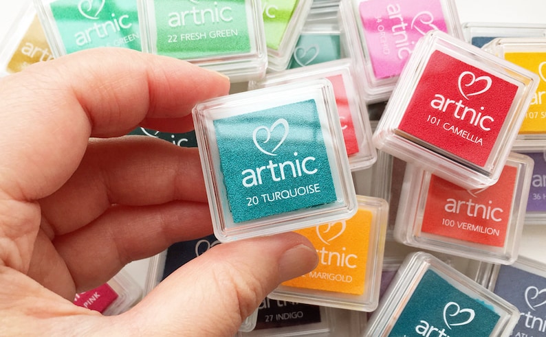Puede incluir: Primer plano de una almohadilla de tinta turquesa etiquetada como "artnic 20 TURQUOISE". La almohadilla de tinta est&aacute; en una caja de pl&aacute;stico transparente y est&aacute; rodeada de otras almohadillas de tinta de varios colores.