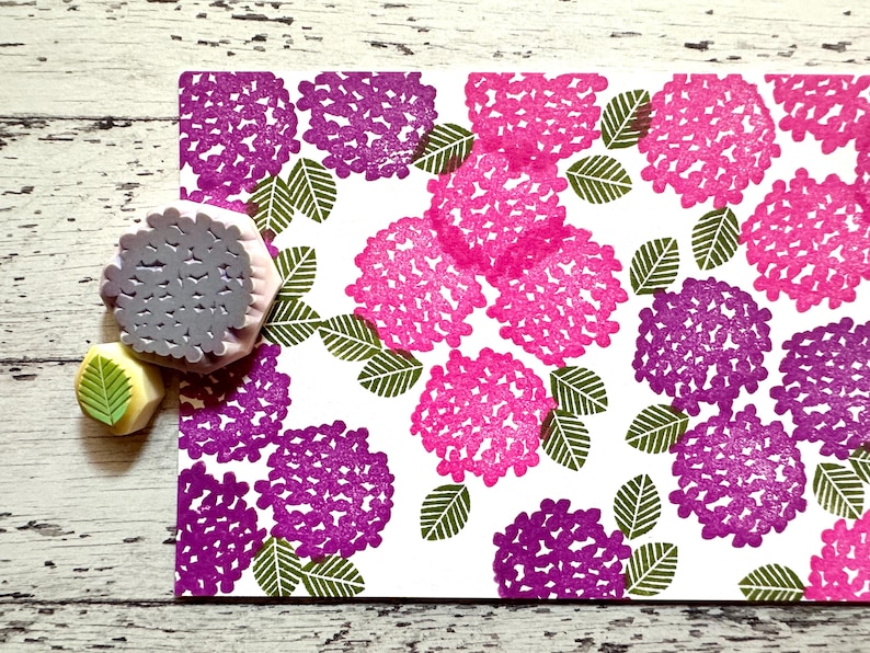 A hand carved rubber stamp set of hydrangea design - flower and leaf, shown with a stamped example in various colored random pattern on white paper.