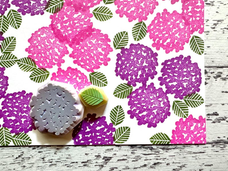 A hand carved rubber stamp set of hydrangea design - flower and leaf, shown with a stamped example in various colored random pattern on white paper.