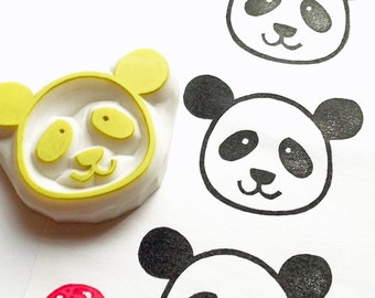 Puede incluir: Un sello de goma blanco con un diseño de cara de panda amarilla. El sello se utiliza para crear impresiones de cara de panda en blanco y negro sobre una superficie blanca. Se muestran tres impresiones de cara de panda.