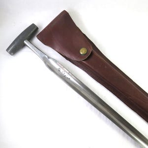 May include: A metal tool with a T-shaped handle and a long, slender shaft, accompanied by a brown leather sheath. The sheath has a snap closure. The tool and sheath are set against a white background.
