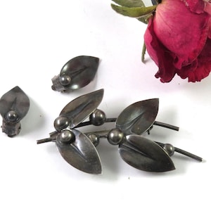 May include: A silver-toned leaf-shaped brooch and matching earrings. The brooch features a series of leaves and small spheres. A dried red rose with green stem and leaves is in the background.