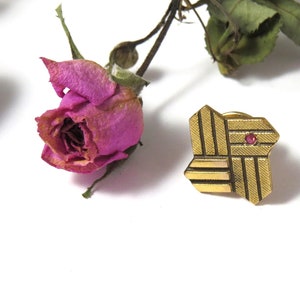 May include: A gold-toned metal pin with a geometric design and a small pink stone in the center. The pin is shaped like a star with six points.