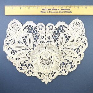 May include: An off-white lace applique featuring an intricate floral and leaf pattern with openwork details. A wooden ruler is positioned above the lace, showing the piece spans approximately 9 inches in width from edge to edge.