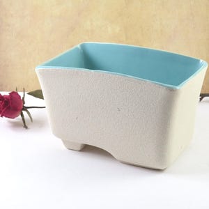 May include: A square, off-white ceramic planter with a textured surface and a light blue interior. The planter has a unique, angled design and sits on small feet. A red rose is visible in the background.