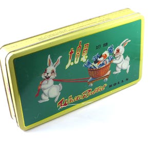 May include: A rectangular yellow tin with a green top featuring a cartoon rabbit design. The design includes two white rabbits, one pulling a cart filled with candy and the other pulling a red ribbon. The text "White Rabbit Rolls" is visible.