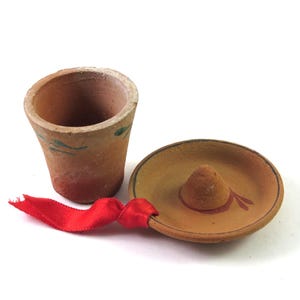 May include: A miniature terracotta shot glass and sombrero set. The shot glass is cylindrical with green painted accents. The sombrero is tan with a brown brim and a red ribbon. The set is likely a decorative item.