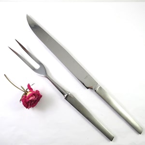 May include: A stainless steel carving knife and a two-pronged carving fork with matte finished handles, positioned diagonally alongside a small, dried pink rose blossom. The knife blade features a small engraved logo near the bolster area.