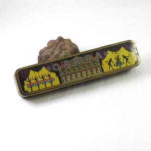 May include: A rectangular vintage tin box with the word "OPERA" in capital letters. The box features illustrations of ballerinas, a theater, and dancers. The background is yellow with a purple curtain design on the ends.