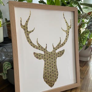 May include: A framed artwork featuring a deer head silhouette made from shotgun shells. The deer head is outlined in white and the background is white.