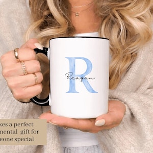 May include: White ceramic mug with a black handle and rim, featuring a large blue letter "R" and the name "Reagan" in script. The mug is held by a person. Text on the image reads "Makes a perfect sentimental gift for someone special."