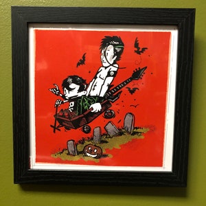 May include: A framed illustration of two cartoon figures in a black wagon flying over a graveyard. The figures are dressed in black and green clothing. The figure in the back is playing a guitar. The wagon is decorated with skulls and crossbones. The background is a red orange color with bats flying around.