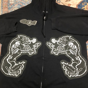 May include: Black zip-up hoodie with two white skeleton wolf patches on the front. The patches are symmetrical and feature the wolves howling with smoke coming from their mouths.