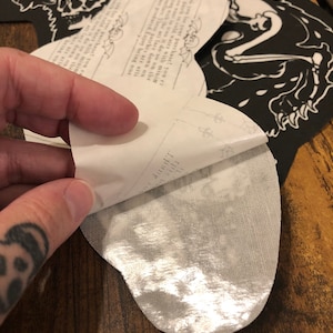 May include: A white iron-on patch with a silver backing. The patch features a black and white illustration of a wolf with a skull and a text that reads "Thank you for your purchase! You can wash the patch in cold water with minimal shrinkage. If you want to iron it, do so on the lowest setting. These patches are artfully produced. Enjoy being one with the art."