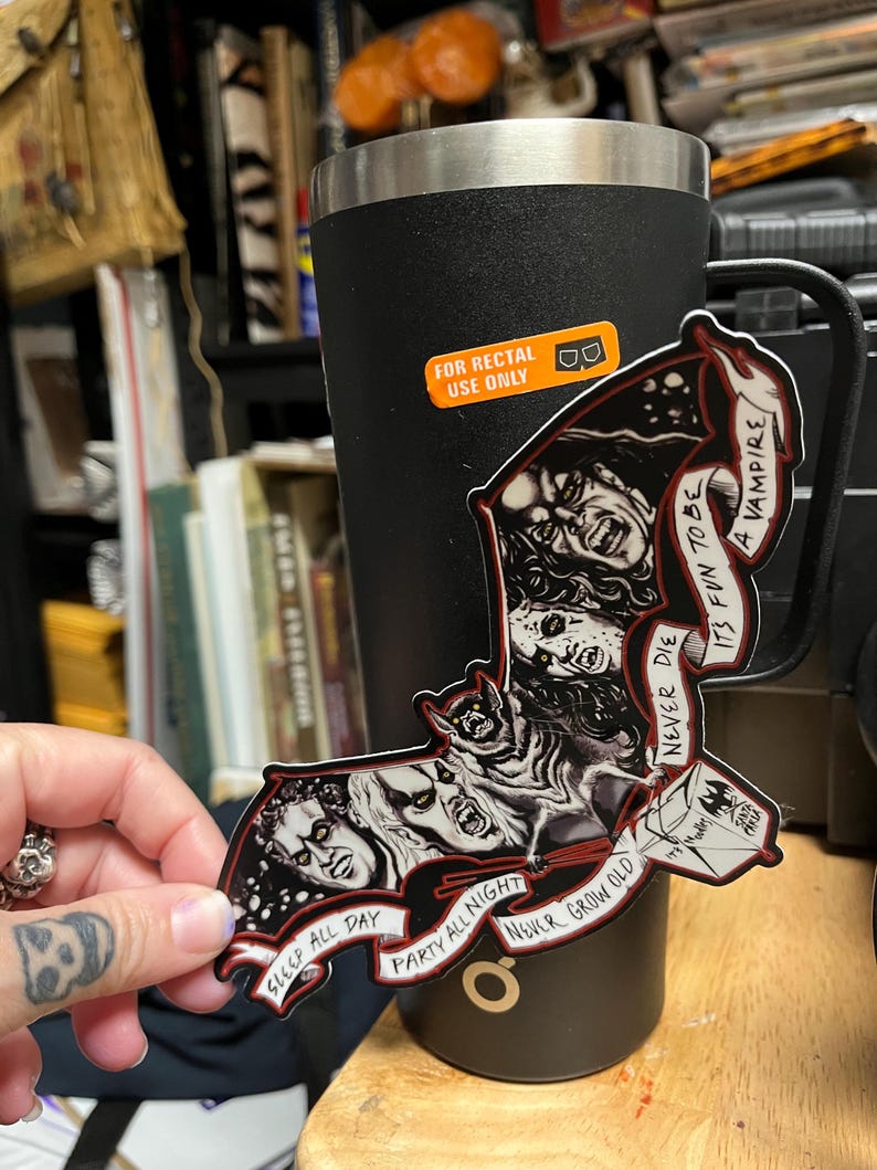 May include: A black and red bat-shaped sticker featuring vampire faces and the phrases "Sleep All Day", "Party All Night", and "Never Grow Old". The sticker is held near a black tumbler with a "For Rectal Use Only" sticker.