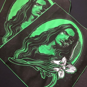 Può includere: Due toppe di tessuto nero con un design verde e bianco. Il design presenta una persona con i capelli lunghi e un fiore. Il viso della persona è parzialmente oscurato da un teschio.