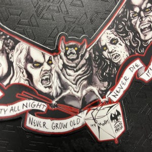 May include: A bat-shaped sticker with a bat holding chopsticks and several vampire faces. The sticker has the text "Party All Night, Never Grow Old" and a Santa Carla Noodles logo.