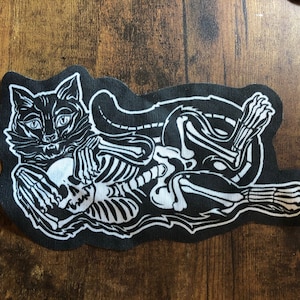 May include: Black and white embroidered patch depicting a cat skeleton holding a skull. The cat is lying on its side with its head resting on the skull. The patch is on a black background.