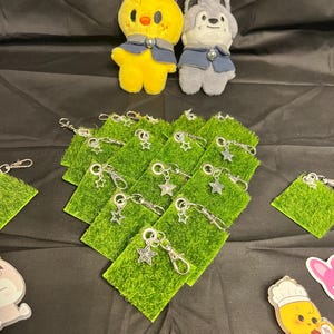 Puede incluir: Un pollito de peluche amarillo y un lobo de peluche gris, ambos con chalecos azules, se encuentran sobre varios llaveros verdes con forma de hierba y colgantes de estrella plateada. Los art&iacute;culos est&aacute;n dispuestos sobre una superficie negra.