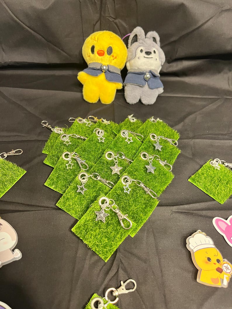 Puede incluir: Un pollito de peluche amarillo y un lobo de peluche gris, ambos con chalecos azules, se encuentran sobre varios llaveros verdes con forma de hierba y colgantes de estrella plateada. Los art&iacute;culos est&aacute;n dispuestos sobre una superficie negra.