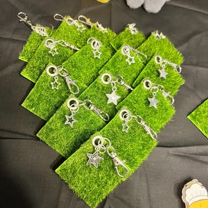 Puede incluir: Llaveros con colgante de estrella plateada exhibidos sobre peque&ntilde;os cuadrados de c&eacute;sped artificial verde. Cada llavero tiene un cierre de mosquet&oacute;n y un dije en forma de estrella. Los llaveros est&aacute;n dispuestos sobre una superficie negra.