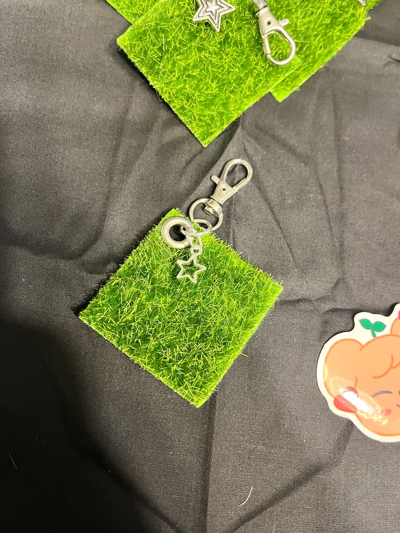 Puede incluir: Un llavero plateado con un dije de estrella adjunto a un peque&ntilde;o cuadrado de c&eacute;sped verde artificial. El llavero tiene un cierre de mosquet&oacute;n para una f&aacute;cil fijaci&oacute;n. Se ven cuadrados de c&eacute;sped y llaveros adicionales.