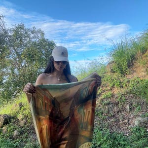 May include: A beige cap and sunglasses are worn by a person holding a scarf. The scarf features a detailed illustration of a European street scene with buildings and the text "LE SAN ASTIA". The background is a bright blue sky and green foliage.