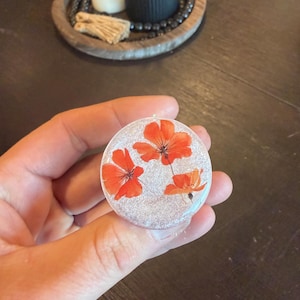 May include: A round, clear resin phone grip featuring three vibrant orange flowers with delicate petals. The grip has a glittery, silver background. The phone accessory is held in a hand.