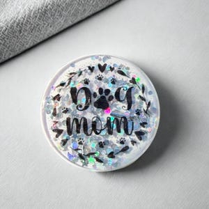 May include: A round, white phone grip with the words "Dog Mom" in black script. The grip is filled with iridescent glitter and small paw prints. Decorative leaves and hearts surround the text.