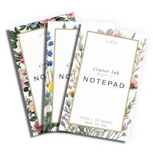 May include: A set of four white notepads with floral borders. Each notepad has a gold border and the text "Crater Ink paper co. NOTEPAD". The notepads are 5x8 inches and have 50 sheets.
