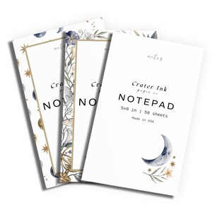 May include: Three white notepads from Crater Ink paper co. measuring 5x8 inches with 50 sheets each. The covers feature watercolor illustrations of moons, stars, and botanical elements with gold-toned frames and the text Crater Ink paper co. NOTEPAD.