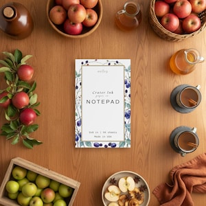 Könnte beinhalten: Ein rechteckiger Notizblock mit weißer Mitte, goldenem Rand und einer botanischen Illustration von Brombeeren und Blättern. Der Text lautet Crater Ink paper co NOTEPAD, 5x8 in, 50 sheets, Made in USA. Der Notizblock ist umgeben von Äpfeln, Tassen und einem Glaskrug.