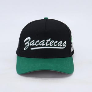 Op de afbeelding: Zwarte en groene baseballpet met het woord "Zacatecas" in wit geborduurd op de voorkant.
