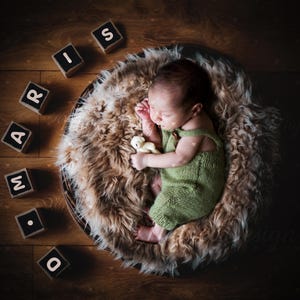 May include: A newborn baby wearing a green knitted outfit lies peacefully in a fluffy, brown fur-lined basket. Wooden blocks spelling out "MARIS" are arranged around the basket on a wooden surface. The baby holds a small stuffed animal.