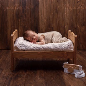 May include: A sleeping baby in a beige knitted outfit lies on a small wooden bed with a white knitted blanket. A wooden toy camera with a white strap rests on the wooden floor. The background is a wooden wall.
