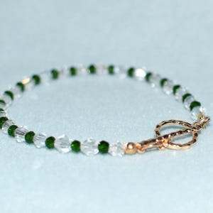 May include: A delicate bracelet featuring clear and emerald green beads. The beads are strung together with a gold-coloured clasp. The bracelet is displayed on a light blue background. This jewellery piece is suitable for everyday wear.