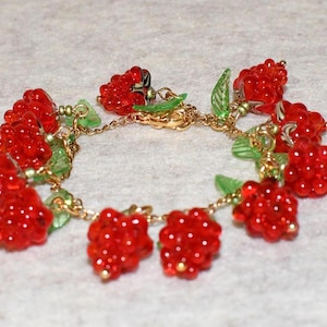 Peut inclure: Un bracelet à chaîne dorée orné de grappes de perles rouges ressemblant à des framboises. Des perles vertes en forme de feuilles et des détails dorés sont intégrés. Un charmant bijou.