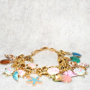 May include: Gold-toned charm bracelet featuring various sea-themed charms. The charms include seashells, starfish, and turtles in shades of blue, pink, green, and orange. The bracelet is displayed against a neutral background.