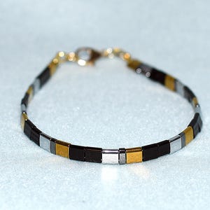 Puede incluir: Una delicada pulsera con pequeñas cuentas cuadradas en colores alternos negro, plateado y dorado. La pulsera tiene un cierre sencillo y está diseñada para llevarse alrededor de la muñeca. Las cuentas miden aproximadamente 0,6 cm.