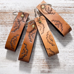 May include: Five brown leather keychains with a geometric pattern and silver key rings. The keychains are arranged in a fan shape on a white wooden surface.