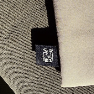 May include: A navy blue label featuring a white kangaroo graphic, sewn onto a textured, dark grey fabric. A light beige fabric is visible on the right. The image shows a close-up.
