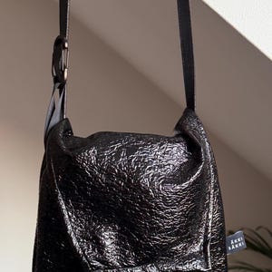 May include: Black shoulder bag with a textured, glossy finish. The bag has a long, thin strap and a small tag with text. The bag is hanging from a strap attached to a metal ring.
