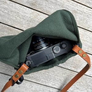 Może przedstawiać: Zielony, skórzany pokrowiec na aparat fotograficzny z brązowym paskiem i mosiężnymi okuciami. Wewnątrz znajduje się czarny aparat z obiektywem.