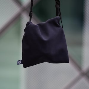 Puede incluir: Un bolso de hombro azul marino oscuro con cierre de cordón. El bolso está hecho de un material suave, posiblemente similar al neopreno. Una pequeña etiqueta rectangular con un gráfico blanco está adherida a la esquina inferior. El bolso está suspendido por una correa negra.