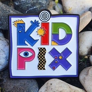 Könnte beinhalten: Eine farbenfrohe Emaille-Anstecknadel mit den Worten "Kid Pix" in einem verspielten Cartoon-Stil. Die Buchstaben sind schwarz umrandet und mit verschiedenen Farben gefüllt, darunter Blau, Rot, Grün, Lila und Gelb. Die Anstecknadel ist auf einem weißen Hintergrund und hat einen schwarzen Rand.
