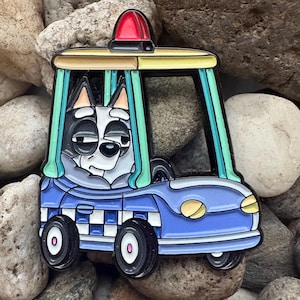 Puede incluir: Un pin de esmalte azul y blanco con un perro de dibujos animados en un coche de policía. El pin tiene una luz roja, detalles amarillos y verdes y ruedas negras. El perro tiene una cara en blanco y negro y lleva un uniforme gris.