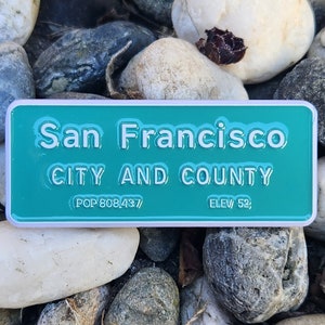 May include: A teal blue enamel pin with white lettering that reads "San Francisco City and County (POP 808,437/ ELEV 52;".