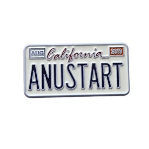 Puede incluir: Un pin blanco con forma de matrícula con el texto "ANUSTART" en letras negritas de color azul oscuro. El pin también presenta la palabra "California" en cursiva, el mes "AUG" y el año "2013". El pin tiene un borde blanco.