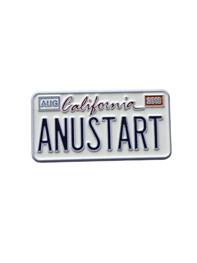 Puede incluir: Un pin blanco con forma de matr&iacute;cula con el texto "ANUSTART" en letras negritas de color azul oscuro. El pin tambi&eacute;n presenta la palabra "California" en cursiva, el mes "AUG" y el a&ntilde;o "2013". El pin tiene un borde blanco.