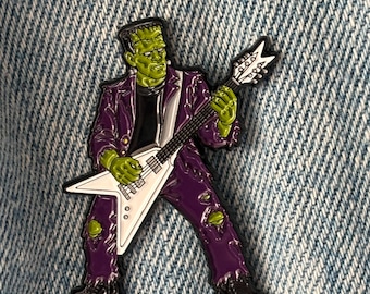 Könnte beinhalten: Ein Metall-Emaille-Pin, der ein grünhäutiges Monster mit Bürstenschnitt zeigt, das einen violetten Anzug und ein schwarzes Hemd trägt und eine weiße V-förmige E-Gitarre spielt, wobei grüne Hände und Knie durch zerrissene Hosen sichtbar sind.
